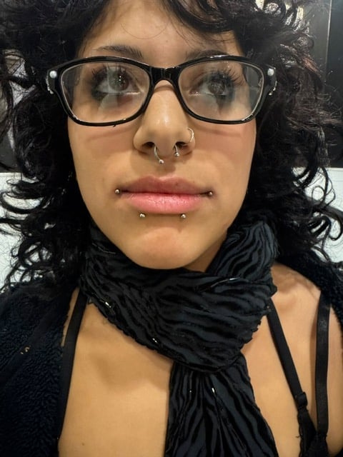 Person with black curly hair and glasses wearing a black scarf around their neck, looking directly at camera