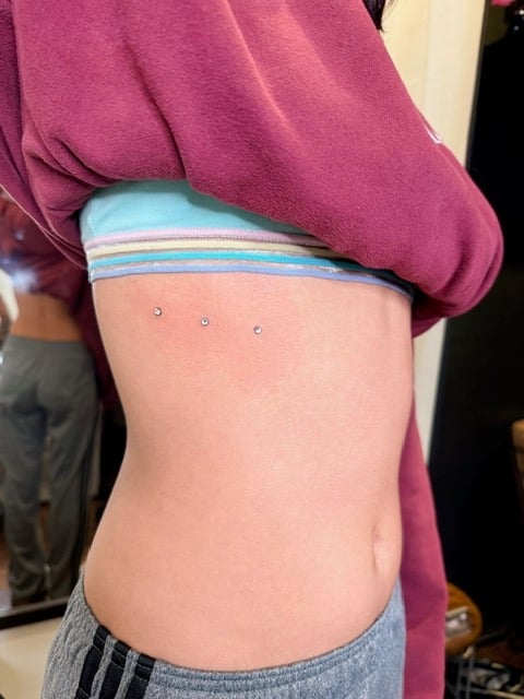 Person showing torso with three dermal piercings on the abdomen, wearing a pink hoodie and blue-striped waistband