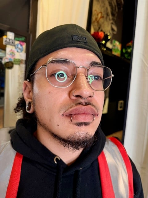 Person wearing black beanie, clear-framed glasses, black and red hoodie with tongue out in playful selfie indoors