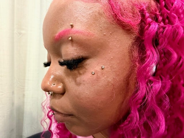 Person with bright pink curled hair, nose and eyebrow piercings, displayed in profile view