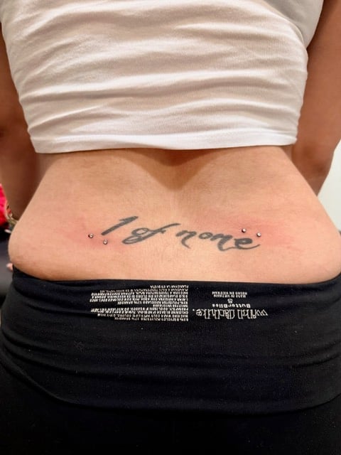 Close-up of a persons lower abdomen showing a cursive script tattoo reading before with two small dots on either side