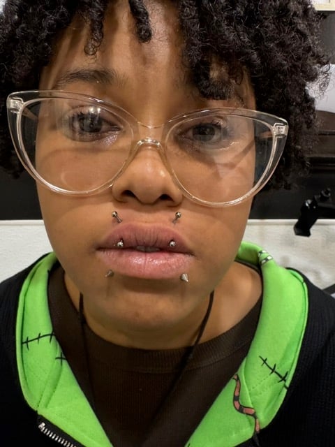 Person wearing clear-framed glasses and facial piercings, with textured black hair, wearing a bright green and black jacket