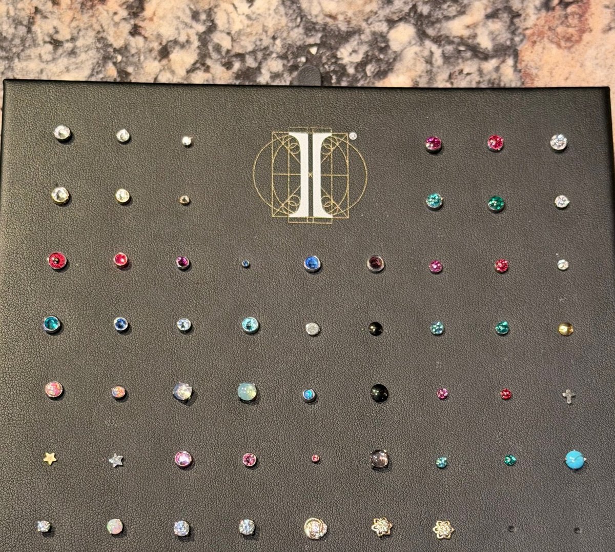 Collection of decorative gemstone studs and earrings in various sizes and colors displayed on a dark sample card with geometric logo