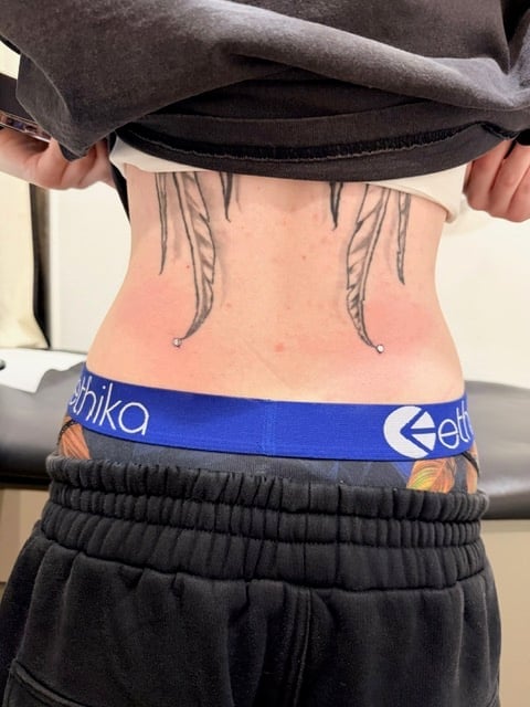 Person showing lower back tattoo of decorative lines, wearing black clothing and blue waistband