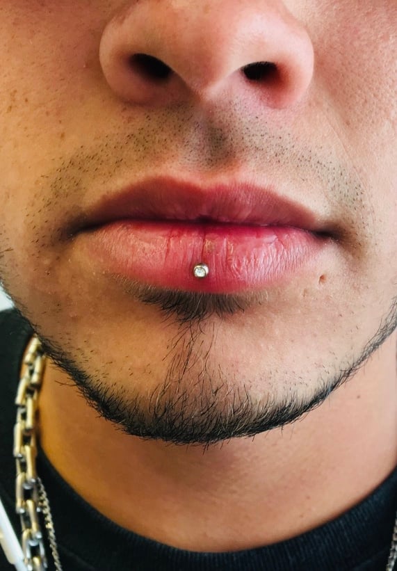 Close-up of a persons face showing a lower lip piercing and facial hair, wearing a chain necklace